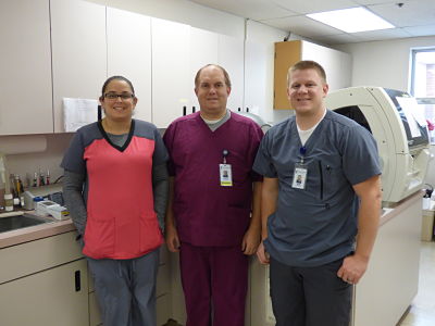 Three Lab works standing next to each other in a Laboratory smiling. There is one female and two males. 