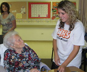 A younger lady with blonde hair is smiling at a older lady sitting in a chair